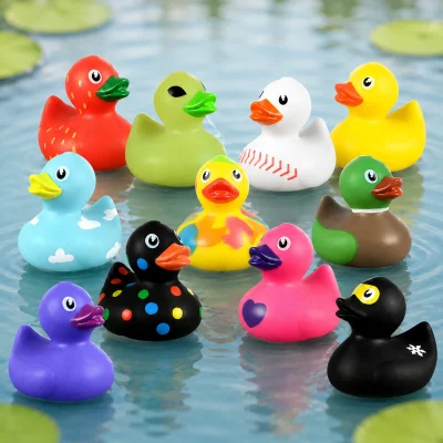 Mini assorted rubber ducks floating on a calm duck pond with soft water ripples and blurred lily pads in the background. Colorful novelty duck designs including strawberry, baseball, cloud, camouflage, polka dot, pink heart, mallard, yellow, purple, and black ducks are grouped together as the focal point.