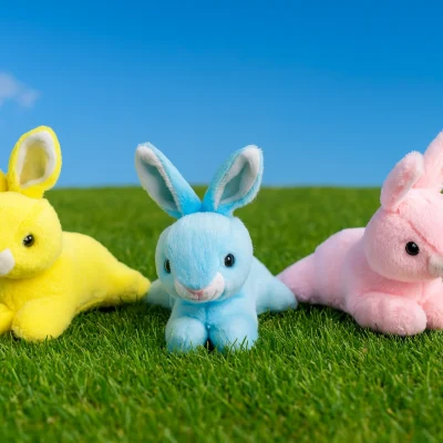 "Three pastel plush bunnies in yellow, blue, and pink sitting on green grass under a clear sunny blue sky with a few clouds."
