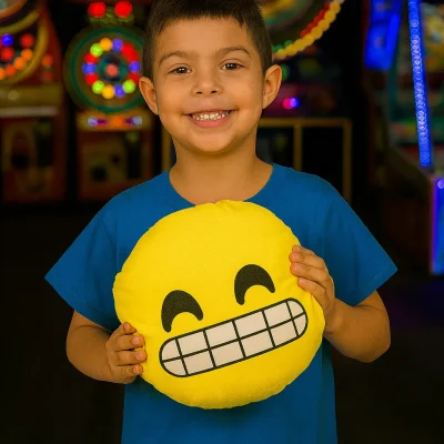 Smiling young boy in a blue shirt holding a yellow emoji pillow with a wide toothy grin, standing inside a brightly lit arcade with colorful game machines in the background.