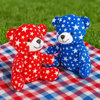 Red and blue patriotic star print stuffed bears sitting on a red, white, and blue picnic blanket on green grass for Fourth of July events