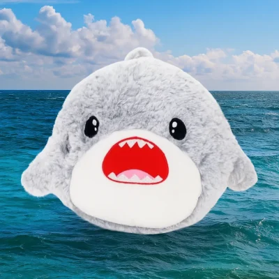A cute gray plush shark toy with a wide open red mouth and cartoonish eyes, floating against a bright blue ocean background with waves and a partly cloudy sky."