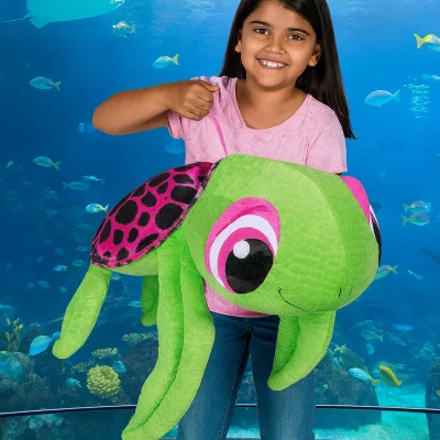 A young girl in a pink shirt smiles while holding a large green plush turtle with pink and black patterned shell, standing in front of a big aquarium filled with fish and a stingray.