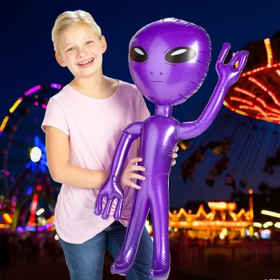 A smiling child holds a large purple inflatable alien at a nighttime carnival, with bright lights, rides, and a Ferris wheel glowing in the background.
