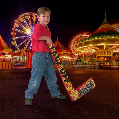 A young boy holding an inflatable hockey stick with flame graphics, standing on a carnival midway at night with bright lights, Ferris wheel, and carousel glowing in the background.