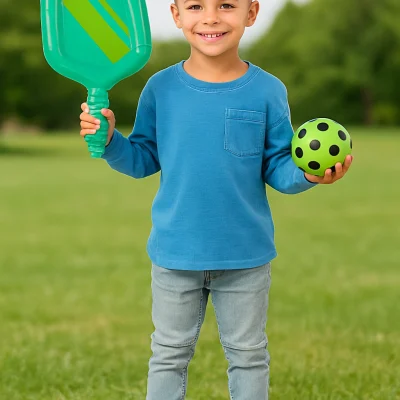 Smiling young boy holding an inflatable paddle and green polka-dot ball while standing on a grassy lawn under a sunny blue sky.