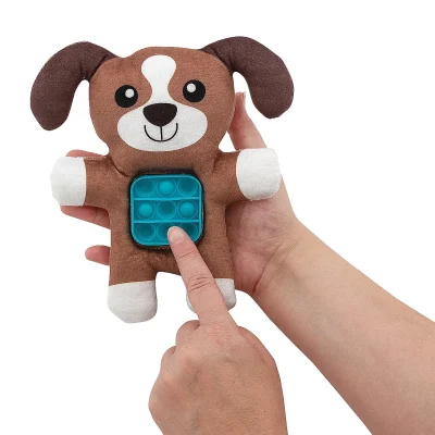 Person holding a brown and white plush dog toy with a blue pop-it fidget panel on its belly, pressing one of the bubbles against a clean white background.